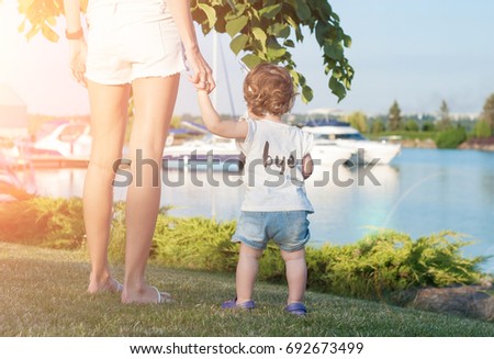 A little girl is holding her mother hand and looking at the yachts on the river