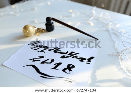 Christmas card with festive inscription Happy New Year, painted with black calligraphic font using carcass and fountain pen, which lies on table with Christmas garland in art workshop. Concept of