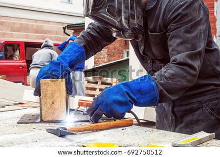Close-up of a man in a black construction uniform, welding mask, protective black gloves, welding a metal yellow construction on a table with a welding machine, a lot of tools around,  red car