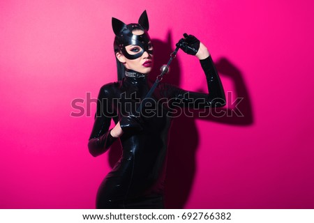 Beautiful dominant brunette vamp mistress girl with fashion makeup in glamour latex dress, collar and bdsm black leather fetish cat mask posing on hot pink backgroung