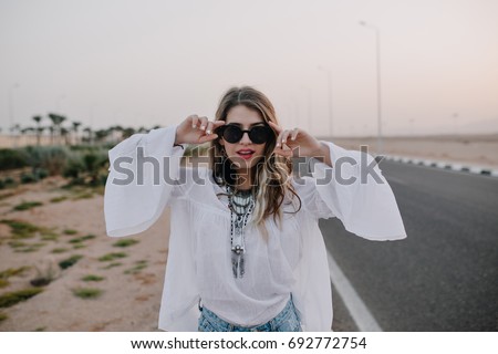 Portrait of graceful stylish girl in black sunglasses and white blouse having fun next to highway in summer. Charming long-haired young woman wearing vintage tunic walking down the road in evening. Royalty-Free Stock Photo #692772754