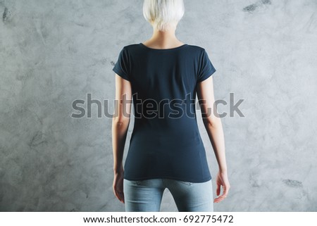 Back view of young woman wearing blank casual dark blue shirt and jeans on concrete wall background. Logo concept. Mock up 