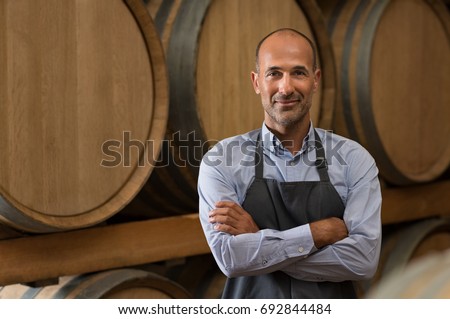 Satisfied winemaker in cellar with folded hand. Happy man standing in his warehouse and looking at camera. Smiling professional man standing in wine cellar with wine wooden barrels in background. Royalty-Free Stock Photo #692844484