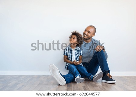 African father and his son sitting on floor and looking up in a blank wall. Happy dad and little boy sitting in an empty room. Young black man with his child thinking and pensive with copy space. Royalty-Free Stock Photo #692844577