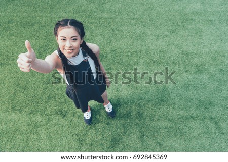 high angle view of smiling asian girl showing thumb up while standing on green lawn 