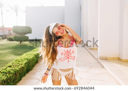 Happy graceful woman with tanned skin dancing and smiling outside on beautiful alley with green bushes. Portrait of lovely girl with dreamy face expression wearing white hat and trendy fringe shirt 