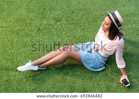 high angle view of smiling african american girl in straw hat listening music with smartphone while sitting on green grass