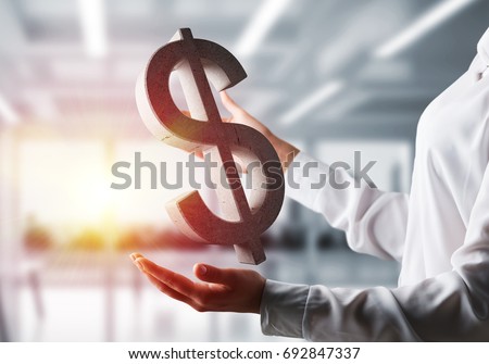 Business woman in white shirt keeping stone dollar sign in hands with office view and sunlight on background. Mixed media.