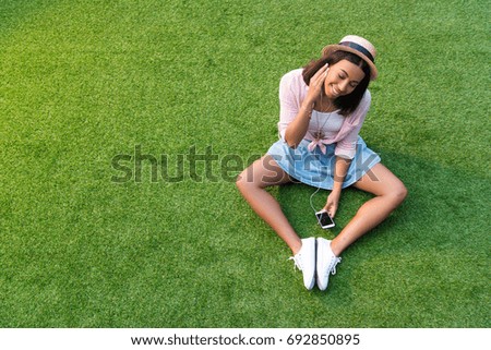 smiling young african american woman in earphones listening music with smartphone while sitting on lawn