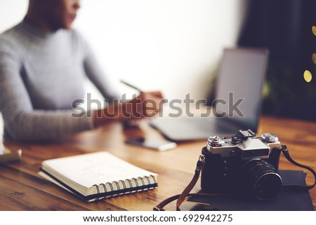 Cropped image of defocused afro american woman freelancer sitting on blurred background editing photographs on laptop computer, selective focus on vintage camera and notebook with blank pages