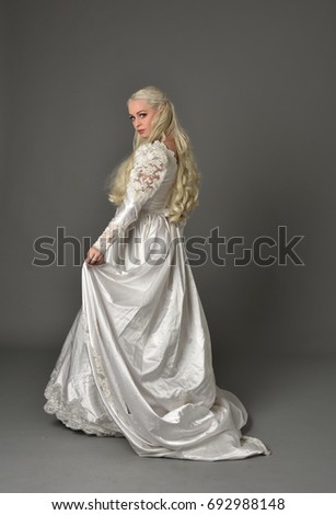 full length portrait of blonde lady wearing bridal gown, standing pose, isolated against a grey background