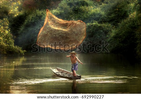 Thai fisherman on wooden boat casting a net for catching freshwater fish in nature river in the early evening before sunset