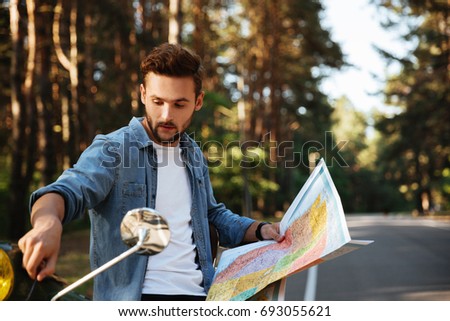 Picture of attractive young bearded man near scooter looking at map outdoors.