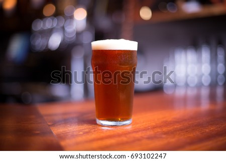Low angle close up perspective of traditional tumbler pint shape beer glass filled with golden malt and hoppy India pale ale with foam head on wood counter top bar with blurry restaurant background Royalty-Free Stock Photo #693102247