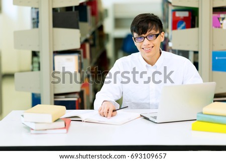 Young student study hard in library. Asian male university student doing study research in library with laptop on desk smiling. For back to school concept and education technology concept. 