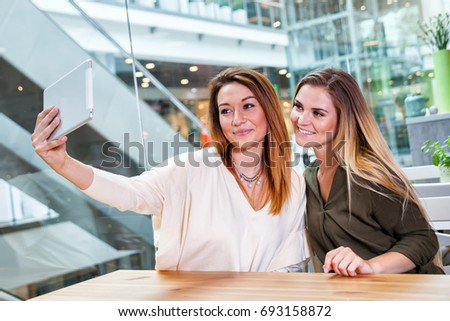 Two woman friends taking selfie with digital tablet in cafe at shopping mall