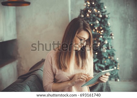Young woman wearing sweater and jeans is buying in Internet with tablet. Christmas tree on background in loft apartment. Christmas and New Year time. Internet shopping