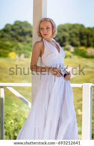 Blond young female in long white dress standing in alcove. Summer day, woman posing for pictures in summer house in meadow.