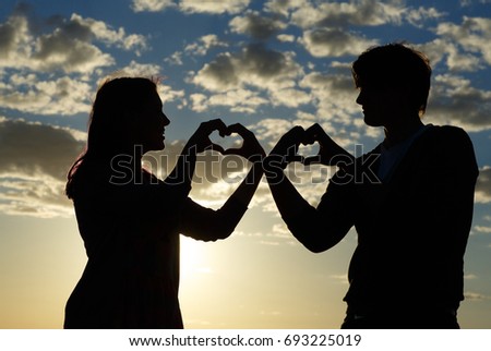 Silhouette of a young girl and her boyfriend with hearts made of fingers against the blue sky with clouds at sunset.  Silhouette of the heart.Lovers in nature.Lovers silhouette.Happy guy and girl


