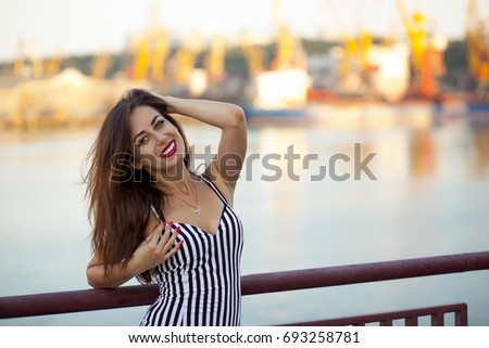 A young smiling beautiful dark-haired woman in a striped black and white dress and red shoes is standing on a bridge near the sea.