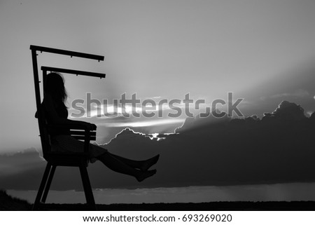 Black and white silhouette of a woman at the beach while sitting on a lifeguard chair in sunset clouds