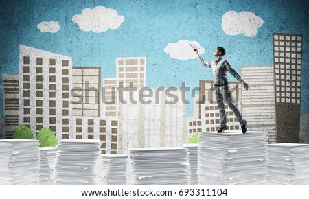 Man in casual wear keeping hand with book up while standing on pile of paper documents with drawn cityscape on background. Mixed media.