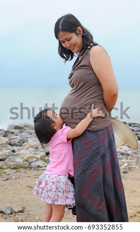 Asian girl gives a lovely kiss at her mother's belly who is pregnant near the seaside,