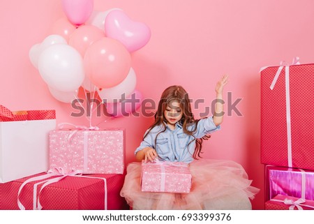 Excited joyful little girl in tulle skirt opening birthday present surround big colorful gift boxes isolated on pink background. Celebrating birthday party with balloons and gifts of amazing kid