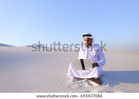 Successful young businessman male Muslim uses laptop to build drawing in Photoshop and prints fingers on laptop sitting on white sand in bottomless wide desert in afternoon against blue sky. Swarthy