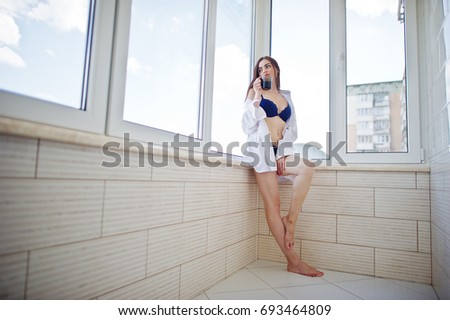 Portrait of a lovely girl in underwear and male shirt standing with a glass of water in her hands on the balcony.
