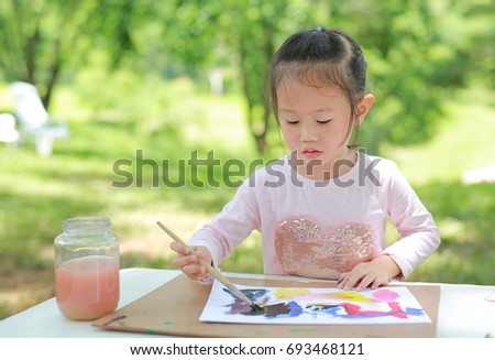 Asian child girl painting with paintbrush on arts paper on the table in the green garden.