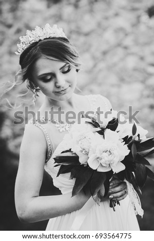 Stylish happy smile bride with bouquet of peonies with crown poses on background in courtyard of house. Close up. Nature. Against the background of the stone wall. Look down. Black and white photo