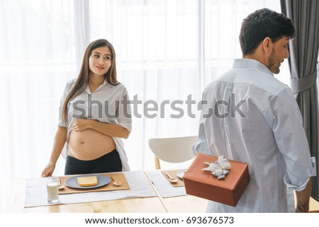 Husband hiding a gift for his wife pregnant waiting in the kitchen room. Concept of family, lover, surprise and birthday.