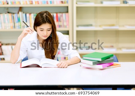 Young student study hard in library. Asian female university student doing study research in library with books on desk and smiling. For back to school education technology concept. Royalty-Free Stock Photo #693754069