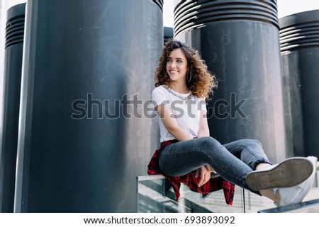 full length picture of pretty stylish girl with wonderful curly hair and open smile in the city. She dressed in white shirt and black jeans. She is looking aside and smiles. Good sunny day. City walks