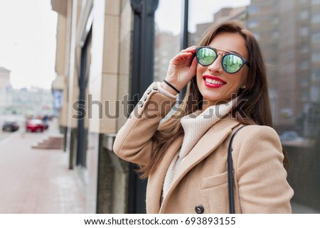 Picture of pretty woman with long dark hair is walking on the street. She has red lips and wearing bright sunglasses. She is looking at camera with charming smile and holds glasses. Street style.