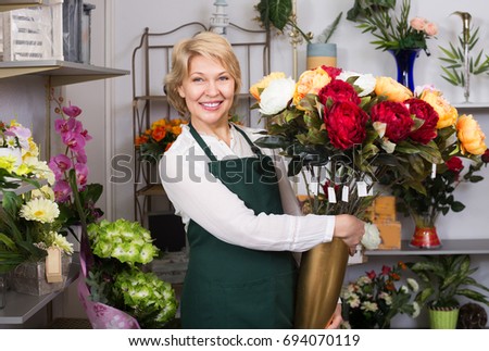 Cheerful mature female florist in apron holding big bouquet with peonies 
