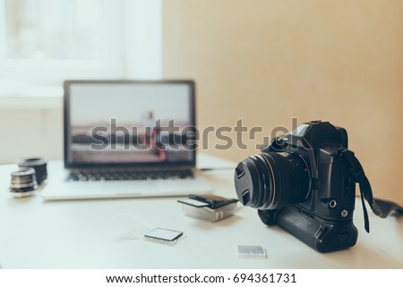 Blur picture of modern laptop with black camera on foreground beside memory cards. Computer stands on table with lens and other accessories.