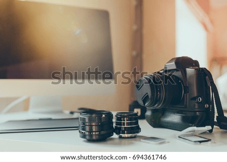 Blur indoor photo of photographer's workplace with camera and lens on table. Close-up picture of modern computer with black screen standing on white desk.
