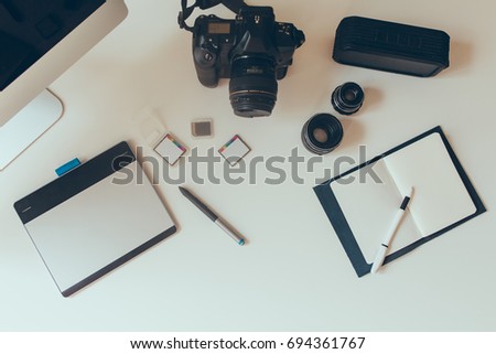 Overhead view of photographer's stuff lie on the white table. Photo from above of black camera, tablet, notebook and pen.