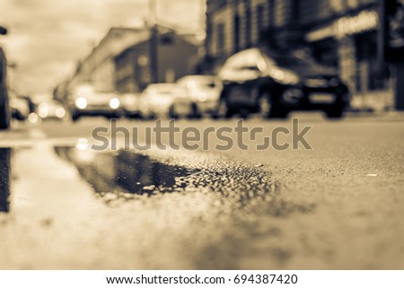 Sunny day after rain in the city, parked cars near the store and the headlights of the approaching cars. Close up view from the level of the puddle on the pavement, image in the yellow toning
