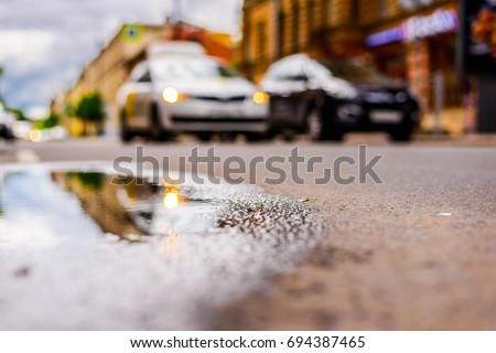Sunny day after rain in the city, parked cars near the store and a passing car. Close up view from the level of the puddle on the pavement