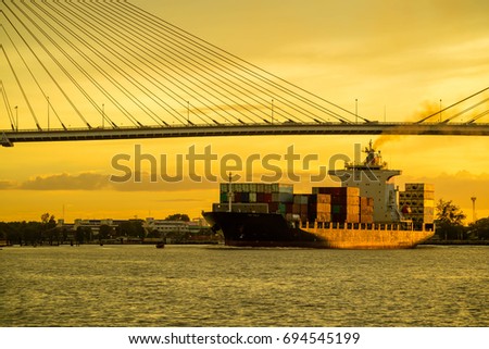 Container cargo ocean ship float on river and pass under bridge at sunset twilight with red sky