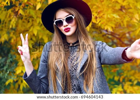 Young stylish woman making selfie in autumn golden leaves park, stylish grunge elegant trendy outfit, dress, gray coat , sunglasses ant vintage hat, making selfie and having fun. Kiss on camera,