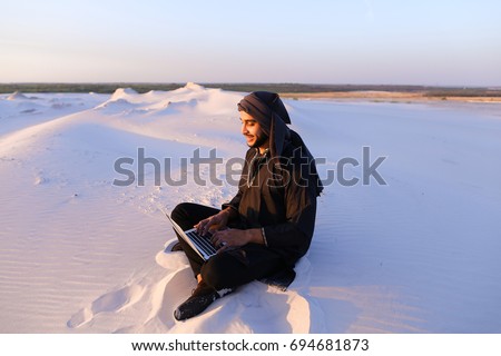 Joyous male Arab sits on sand at computer. young man engaged in scientific work or writes article typing on keyboard of gadget, relaxes sitting in silence of bottomless desert against blue sky on