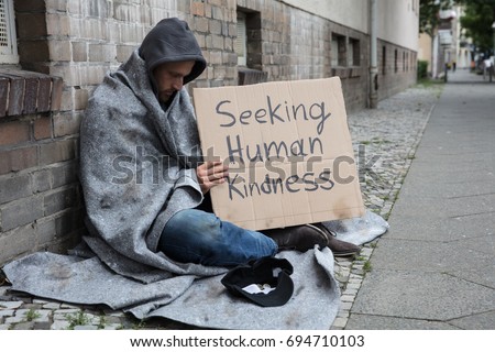 Male Beggar In Hood Showing Seeking Human Kindness Sign On Cardboard Royalty-Free Stock Photo #694710103