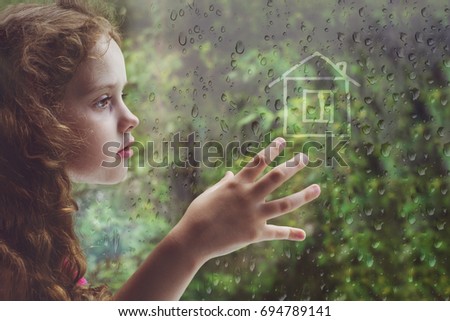 Sad curly little girl is drawing house on the raindrop window.