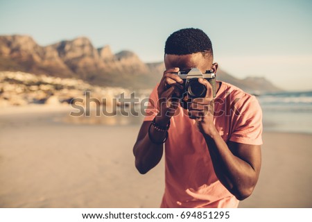 Black guy using digital camera on beach. Afro american man taking pictures on the seashore.