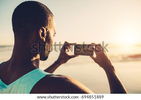 Rear view of young man taking picture of scenery with mobile phone. African man doing mobile photography on the beach.
