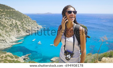 A female tourist photographer speaks on the phone, with a backpack on her back and wearing sunglasses, a wonderfully beautiful view of the blue sea and mountains. Concept: communication, travel, call
